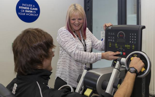 A member of staff assisting a gym member with an arm bike